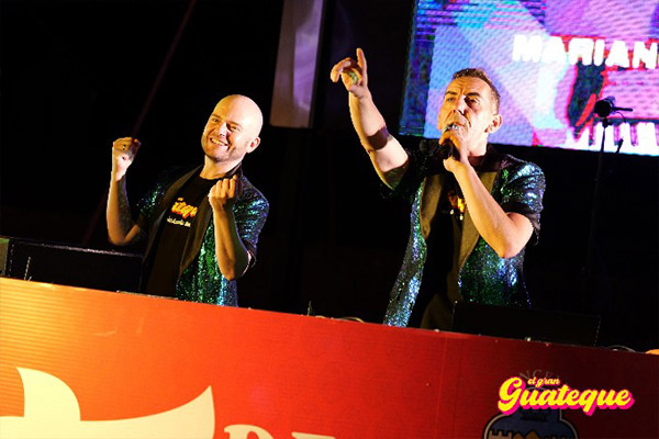 X.avi PetardPop y Edi FM en la cabina de Video DJs, sonrientes y saludando al p�blico; Eddy con micr�fono se�alando y X.avi con las manos levantadas, ambos con chaquetas verdes de brillantina y camisas negras del Gran Guateque; fondo con pantalla y p�blico animado